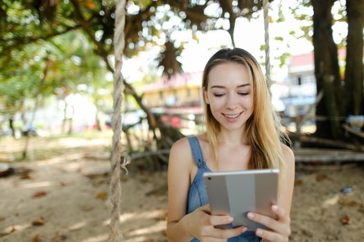 Young nice girl using tablet and riding swing on sand, wearing jeans sundress. Concept of summer vacations and modern technology.