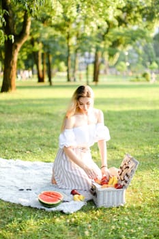 Young woman preparing for picnic in park, sitting on plaid near box and waterlemon. Concept of having leisure time, resting in open air and summer season.