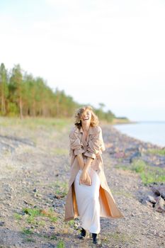 Young girl wearing coat and white dress standing on sea beach with trees in background. Concept of summer vacations photo session and freedom, happiness.