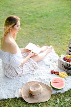 Young caucasian girl reading book and sitting on plaid near fruits in park. Concept of having summer picnic and leisure time.