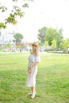 Young girl wearing hat and dress, standing in park on grass. Concept of summer season fashion and female person.