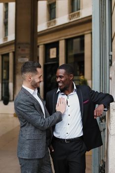 Two happy men, caucasian and afro american, wearing suits speaking near building outdoors. Concept of successful business partners.