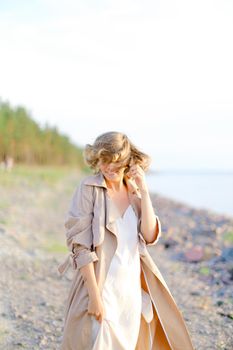 Young smiling woman standing on sea beach and wearing coat with white dress. Concept of enjoying freedom and summer photo session.