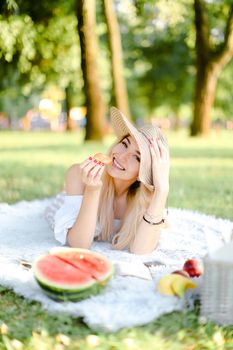 Young happy blonde woman in hat with croissant lying in park on plaid near waterlemon. Concept of having free time, picnic and bakery products.