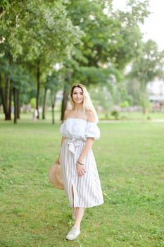 Young blonde girl wearing dress, standing in park and keeping hat. Concept of summer season and fashion.