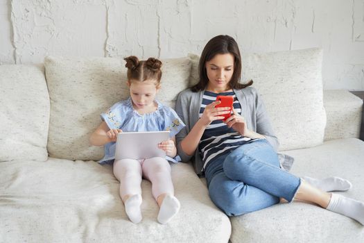 Mother and daughter using electronic devices sitting on sofa at living room