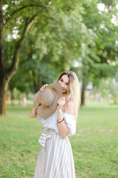 Young jocund girl wearing dress, standing in park and keeping hat. Concept of summer season and fashion.