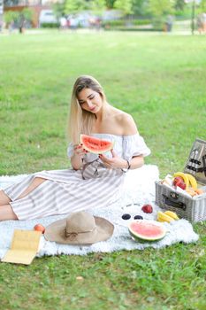 Girl sitting on plaid near fruits and hat, eating watermelon, grass in background. Concept of summer picnic and resting on weekends in open air.