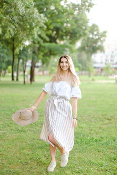 Young girl wearing dress, standing in park and keeping hat. Concept of summer season and fashion.