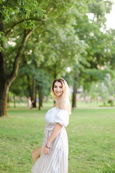 Young pretty girl wearing dress, standing in park and keeping hat. Concept of summer season and fashion.