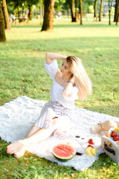 Young caucasian woman sitting on plaid with fruits in park. Concept of summer picnic, photo session in open air and vacations.