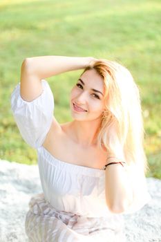 Portrait of young blonde smiling woman sitting on plaid, grass in background. Concept of beautiful female person and summer season.