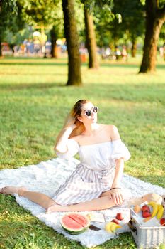 Young caucasian lady in sunglasses having picnic on plaid and sitting in park with fruits. Concept of resting in open air, leisure time and summer season