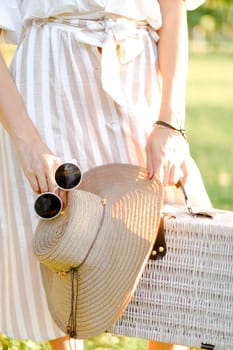 Close up bag, hat and sunglasses in female hands, dress in background. Concept of summer fashion.