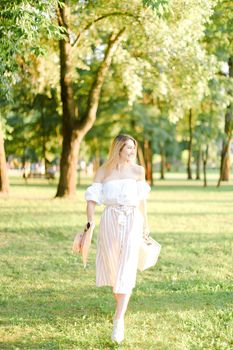 Young blonde girl walking in garden and keeping bag, sunglasses and hat. Concept of walking in park and summer fashion.