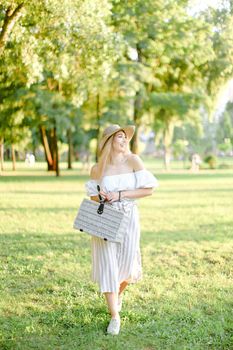 Young charming girl walking in garden and keeping bag, sunglasses and hat. Concept of walking in park and summer fashion.