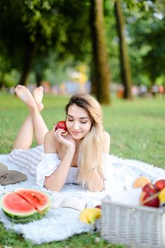 Young nice woman lying on plaid in park, reading book near watermelon and keeping apple. Concept of leisure time, summer vacations and picnic, healthy food.