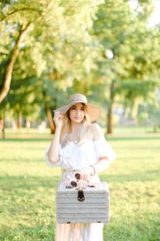 Young nice woman wearing hat and dress standing in garden with bag. Concept of beautiful female person, summer fashion and walking in park.