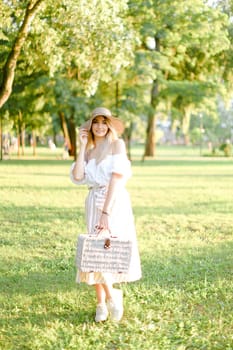 Young blonde woman wearing hat and dress standing in garden with bag. Concept of beautiful female person, summer fashion and walking in park.