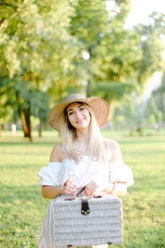 Young woman wearing hat and dress standing in garden with bag. Concept of beautiful female person, summer fashion and walking in park.