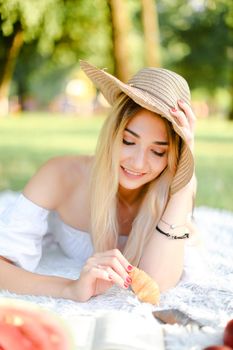 Young caucasian woman in hat lying in park on plaid and surfing by smartphone. Concept of fast internet, picnic and modern technology, bakery products.