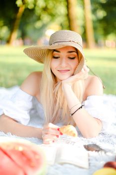 Young blonde woman in hat keeping croissant, lying in park on plaid and reading novel. Concept of picnic and interesting book, bakery products.