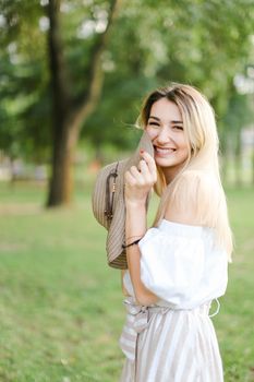 Young beautiful girl wearing dress, standing in park and keeping hat. Concept of summer season and fashion.