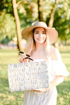 Young pretty woman wearing hat and dress standing in garden with bag. Concept of beautiful female person, summer fashion and walking in park.