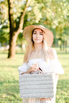 Young charming woman wearing hat and dress standing in garden with bag. Concept of beautiful female person, summer fashion and walking in park.