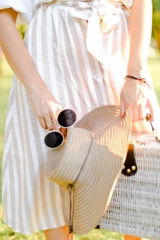 Close up bag, hat and black sunglasses in female hands, dress in background. Concept of summer fashion.