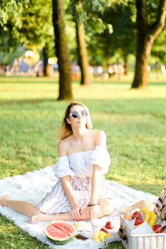 Young blonde caucasian girl in sunglasses having picnic on plaid and sitting in park with fruits. Concept of resting in open air, leisure time and summer season