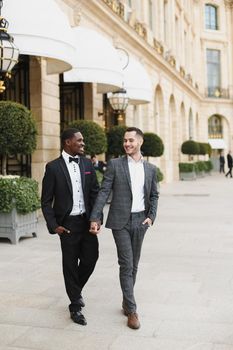 Afro american and caucasian happy handsome gays walking outside and holding hands in city. Concept of same sex male couple.