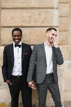 Afro american and caucasian smiling gays standing near building and wearing suits. Concept of lgbt and walking in city.