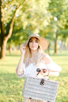 Young happy woman wearing hat and dress standing in garden with bag. Concept of beautiful female person, summer fashion and walking in park.