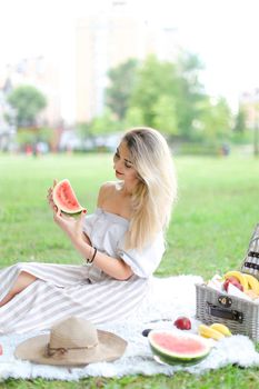 Young girl eating watermelon, sitting on plaid near fruits and hat with grass in background. Concept of summer picnic and resting on weekends in open air.