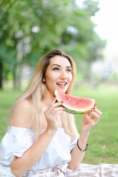 Young woman eating watermelon and wearing dress, grass in background. Concept of sumamer photo session, picnic.