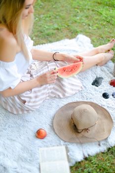 Young blonde girl sitting on plaid near fruits and hat, eating watermelon, grass in background. Concept of summer picnic and resting on weekends in open air.