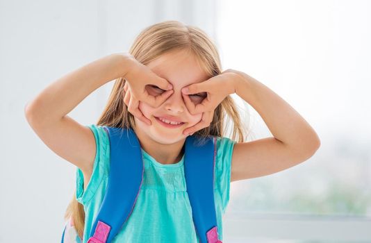 Little girl with backpack makes glasses with her hands.