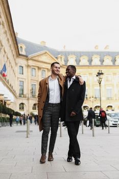 Caucasian smiling man in suit walking with afroamerican male person and hugging in city. Concept of happy lgbt gay and strolling.