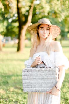 Young cute woman wearing hat and dress standing in garden with bag. Concept of beautiful female person, summer fashion and walking in park.