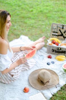 Young blonde female person sitting on plaid near fruits and hat, eating watermelon, grass in background. Concept of summer picnic and resting on weekends in open air.