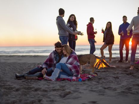 Young Couple Sitting with friends Around Campfire on The Beach At sunset drinking beer