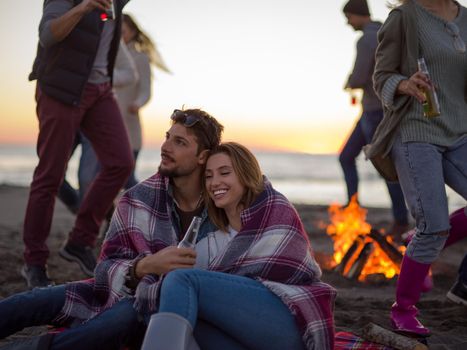 Young Couple Sitting with friends Around Campfire on The Beach At sunset drinking beer