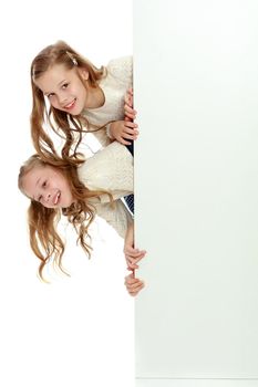 Two cute little girls look out from behind a white advertising banner. On the banner you can make any advertising inscription or put a picture or logo. It is isolated on a white background.