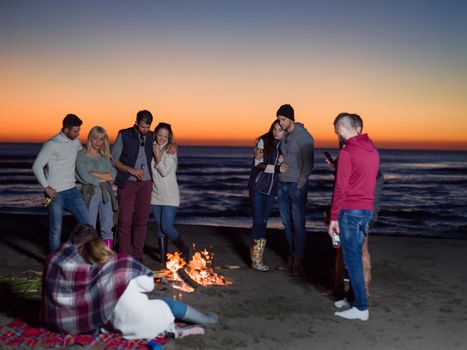 Happy Carefree Young Friends Having Fun And Drinking Beer By Bonefire On The Beach As The Sun Begins To Set