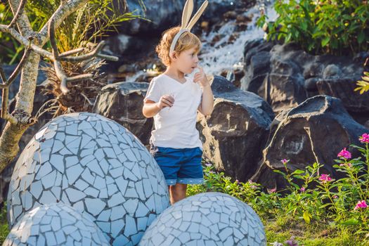 Cute little kid boy with bunny ears having fun with traditional Easter eggs hunt, outdoors. Celebrating Easter holiday. Toddler finding, colorful eggs.