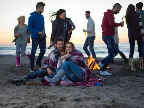 Young Couple Sitting with friends Around Campfire on The Beach At sunset drinking beer