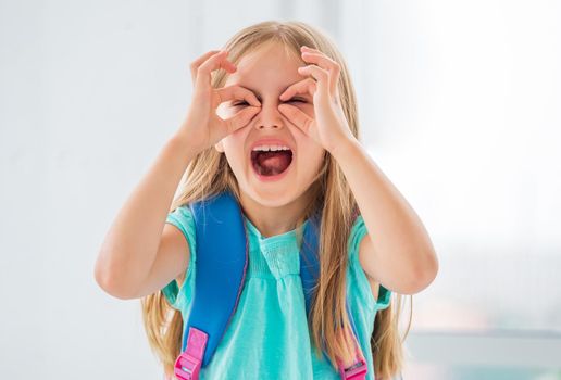 Laughing little girl with backpack fooling making glasses with her hands.