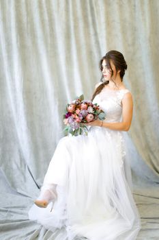 Caucasian brunette bride sitting at photo studio with bouquet of flowers. Concept of wedding and fianceee fashion.