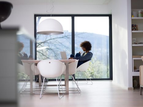 Young african american woman smiling sitting near bright window while looking at open laptop computer on table and holding white mug in her luxury home